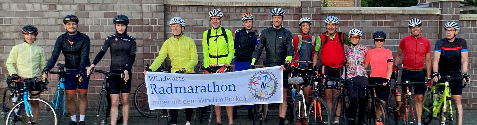Eine Gruppe Radfahrer*innen stehen vor einem Gebäude und halten ein Banner hoch. Darauf steht: Windwärt Radmarathon - Immer mit dem Wind im Rücken