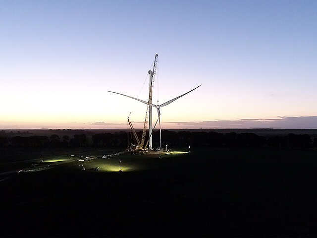 Eine Baustelle für eine Windenergie-Anlage. Es dämmert bereits. Am Horizont ist erkennbar, wie ein Baukran die Rotorblätter zur Turbine hochzieht.