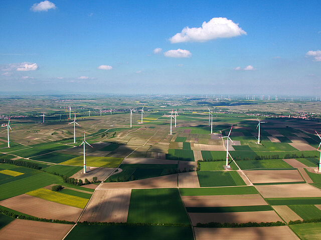 Der Windpark Gau-Bickelheim aus der Vogelperspektive. Die Anlagen stehen in Mitten von Feldern. Ringsum sind Gemeinden zu erkennen. Der Himmel ist blau und nur vereinzelt bewölkt.