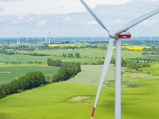 Eine Windkraft-Anlage aus der Vogelperspektive. Die Landschaft im Hintergrund grünt. Am Horizont stehen weitere Windkraft-Anlagen, sowie gelbe Rapsfelder.