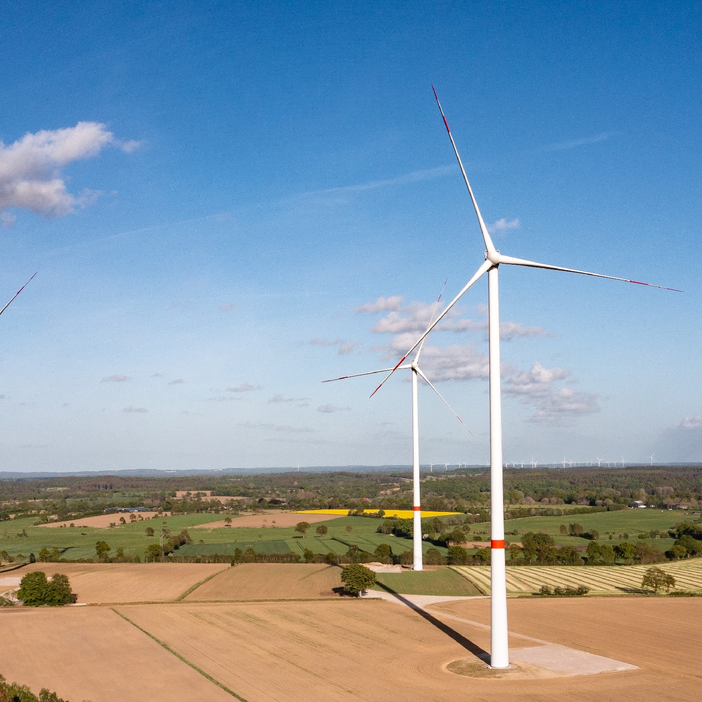 Mehrere Anlagen des Windparks Thaden, die auf landwirtschaftlich genutzten Flächen stehen. Die Sonne scheint, der Himmel ist blau mit einigen kleinen Wölkchen